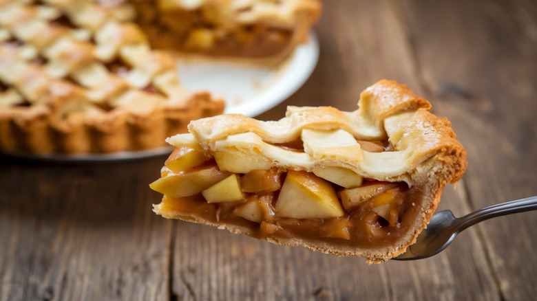 Slice of apple pie on a fork with a whole apple pie in the background.
