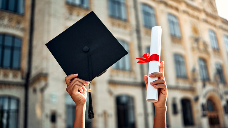 Someone holding up a diploma and a graduating cap in front of an old university building.