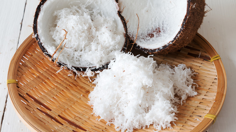 Close-up shot of fresh grated coconut flesh and coconut shell on bamboo tray.