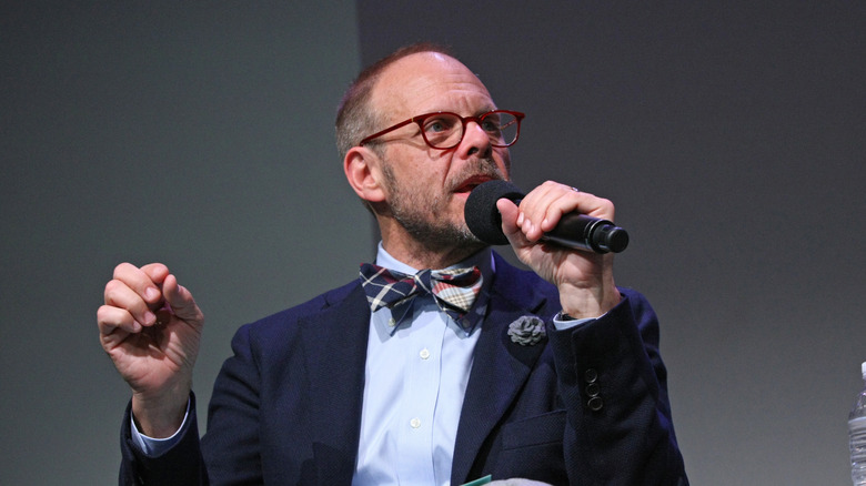 Alton Brown sitting on stage talking into a microphone at an event with a bowtie on.