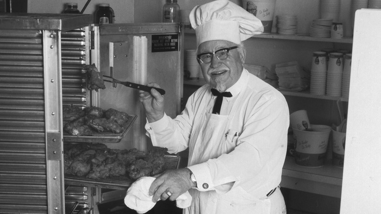 Colonel Harland Sanders, wearing a chef's hat, holds a piece of chicken with a kitchen fork