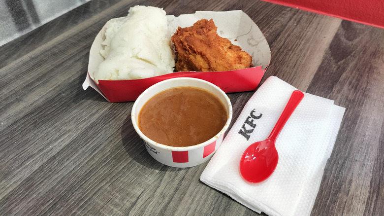 A container of KFC gravy rests next to fried chicken, mashed potatoes, a napkin, and fork on a wood-patterned surface