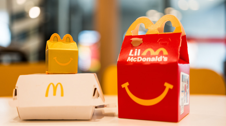 A toy of the Gold Happy Meal Box from McDonald's, next to a Lil McDonald's container and white McDonald's takeout box.
