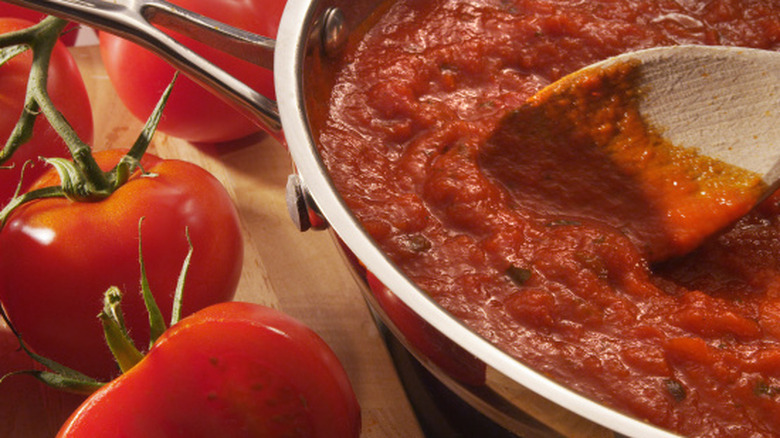 A wooden spoon stirring fresh marinara sauce in a pan beside several tomatoes on a wooden table beside it