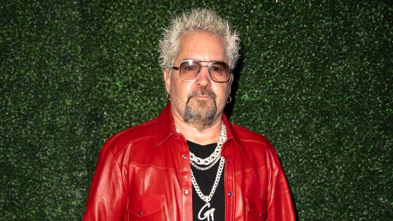 Guy Fieri wearing sunglasses, a red jacket, and a large G chain around his neck in front of a green hedge