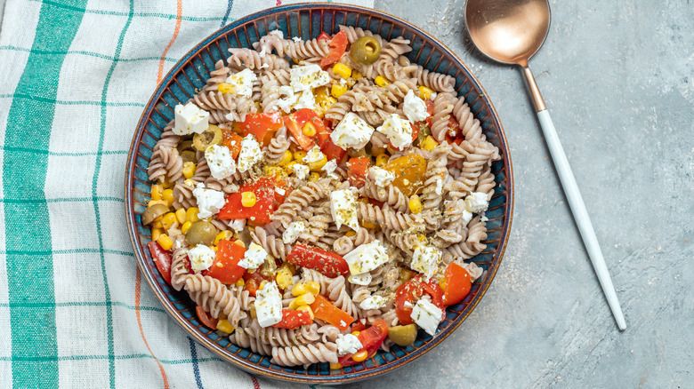 Aerial view of pasta salad served in a bowl with a large spoon, and a napkin on a concrete table.