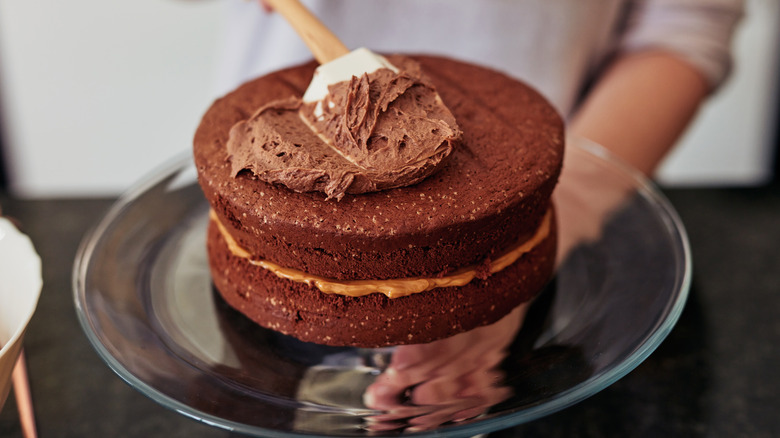 Hands frosting a chocolate cake on a cake stand