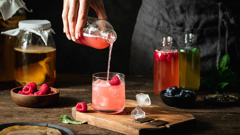 Hand pouring raspberry kombucha into glass with kombucha bottles and jar with scoby in background