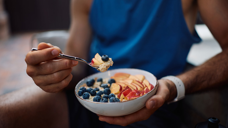 person wearing blue exercise shirt holding bowl of oatmeal with blueberries and peaches