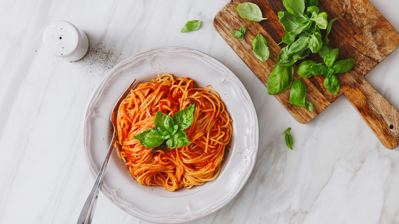 Aerial view of spaghetti and sauce on a white plate with a fork, and a cutting board with leaves on the upper righthand side, and a pepper shaker to the left, all on a marble table.