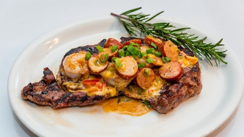 a ribeye steak topped with a Cajun cream sauce with smoked sausage, shrimp, and scallions on a white plate