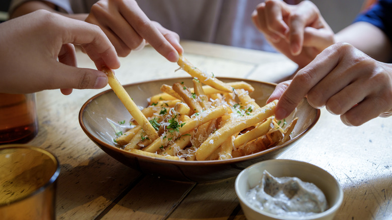 Group of hands sharing french fries with white dipping sauce on the bottom righthand side, all on a wooden table.