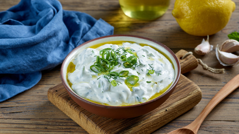 Bowl of sour cream with olive oil and chopped chives on top, with a lemon, garlic cloves, a wooden spoon, and a blue napkin behind.