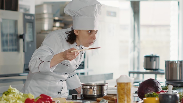 Chef wearing uniform holding utensil near mouth over a pot on the hob with a surprised expression