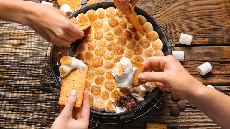 Four hands enjoying a bowl of s'mores dip on a wooden table
