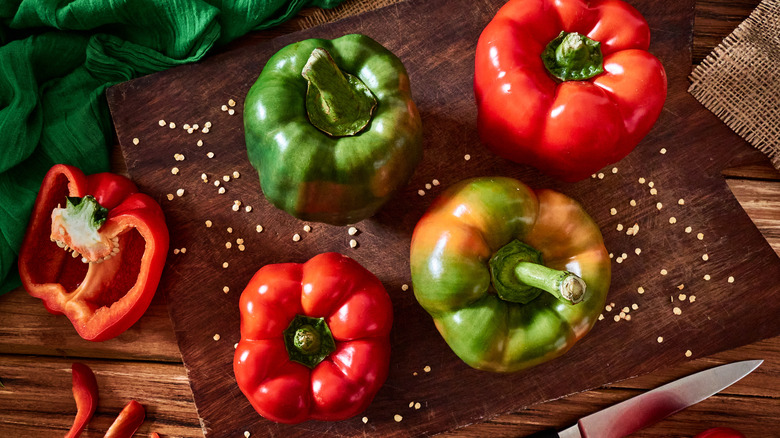 Green and red bell peppers sitting on a cutting board with pepper seeds around them