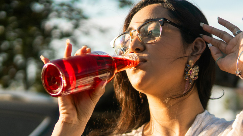 A woman drinks red soda from a glass bottle