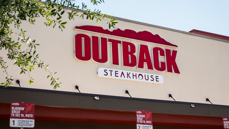Front of an Outback Steakhouse showing name in red and white on a cream-colored facade