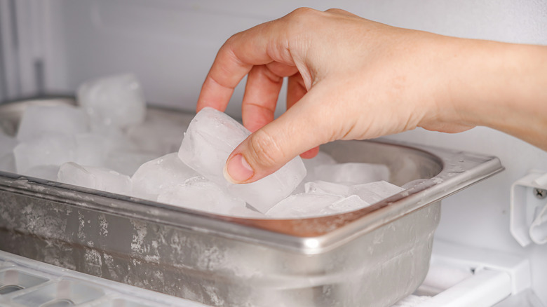 A hand picks up an ice cube from a metal bin in a freezer