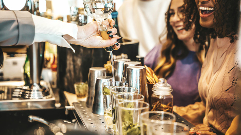 Two women smiling as a bartender pours drinks in front of them.