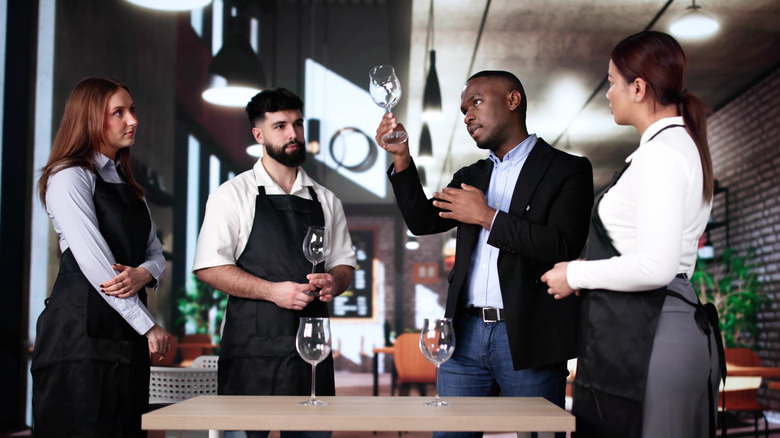 Man in blazer and shirt holding drinking glass in the air among three people wearing aprons in restaurant