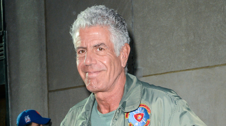Anthony Bourdain smiling as he walks outside in a green bomber jacket.