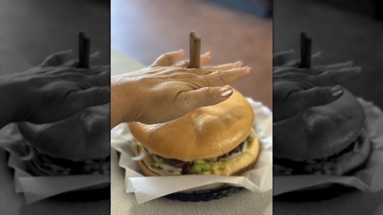 A person holding their hand over a hamburger with a knife stuck in the center in a paper-lined basket to show that the burger is as large as their hand