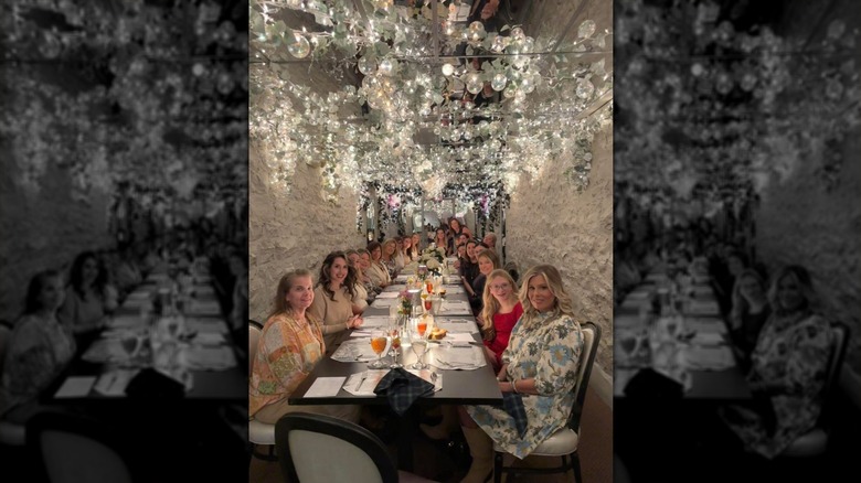 The side view of a very long table in a narrow room with opulent silver bauble decorations hanging from the ceiling; approximately 10 women are seated at each side of the table smiling at the camera