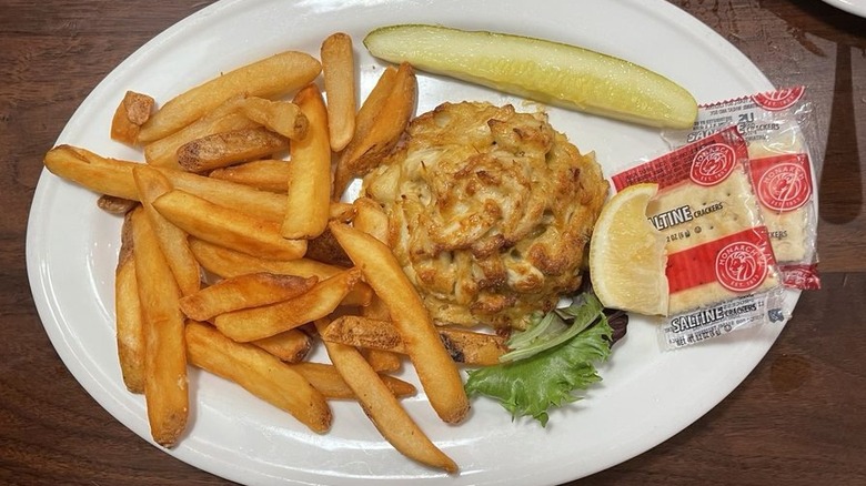 A large crab cake on oval white plate with pickle spear, fries, and 2 cracker packets