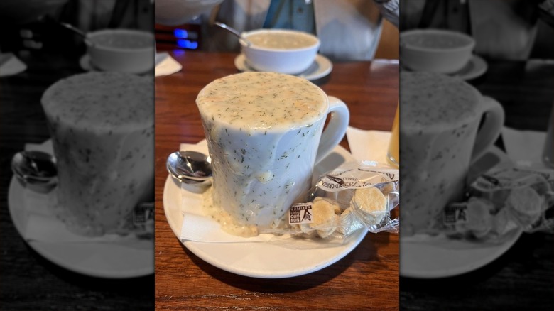 A white mug  on a white saucer overflowing with clam chowder with a package of oyster crackers
