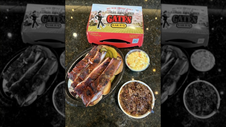 Ribs on top of two pieces of white bread and pickles on a black plate with potato salad and burnt ends and a Gates Bar-B-Q to-go box