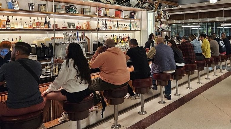 People seated on backless stools at a long counter with a fancy back bar stocked with booze bottles behind it