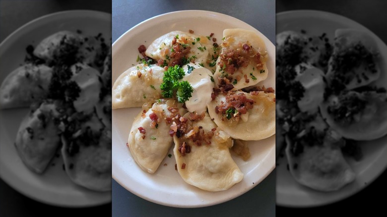 Six pierogi arranged on a white plate topped with bacon, sour cream, and herbs