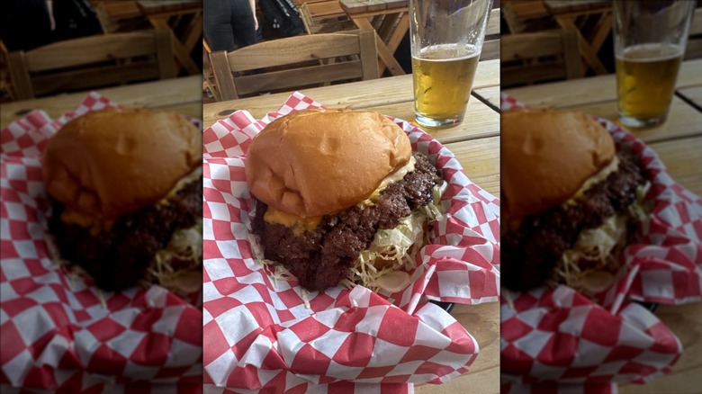 A cheeseburger in a red-and-white checkerboard paper-lined basket with half a pint of beer in the background