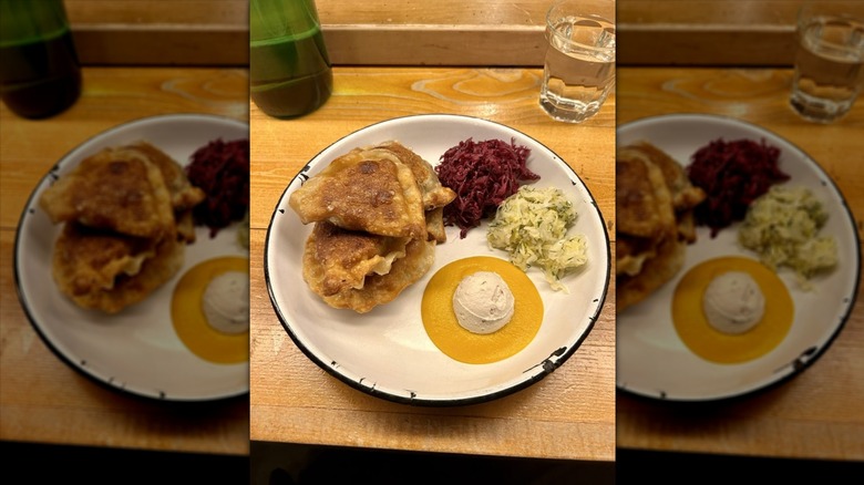 A white plate on a wood surface with four pierogi, shredded red and green cabbage, sour cream, and mustard