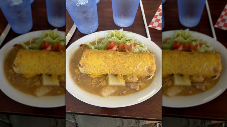 A white plate with a cheese-covered burrito with green sauce, lettuce, and tomato