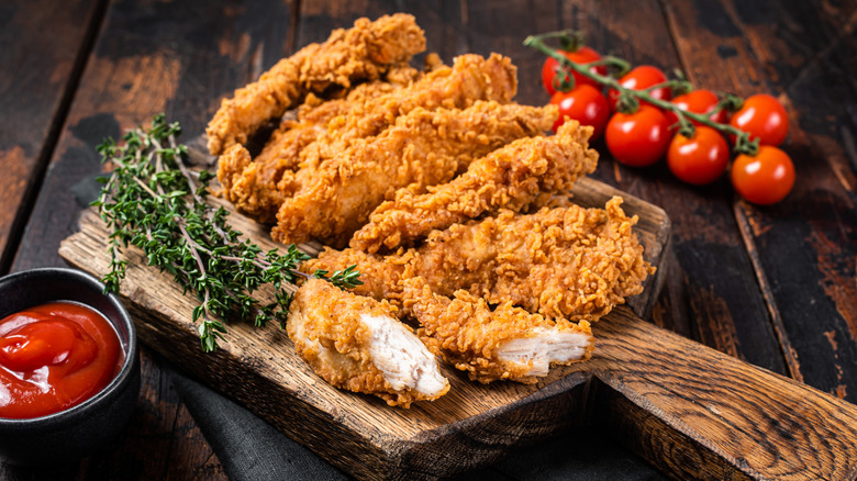Crispy chicken tenders on a wooden board, with herbs, ketchup, and fresh tomatoes