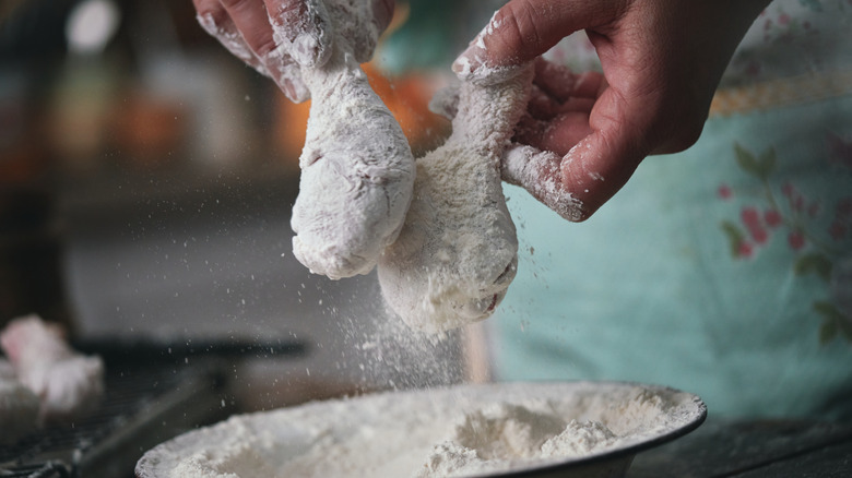 A person dips chicken into a bowl of flour in a kitchen
