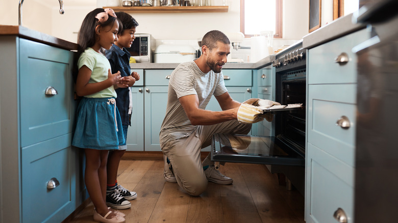 A man kneels to place a baking sheet in an oven while two children stand nearby