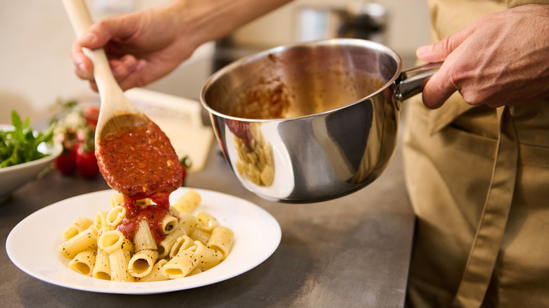 A person in an apron spooning tomato sauce over a dish of herbed pasta