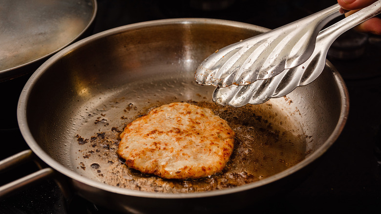 A pair of silver tongs reaches for a turkey burger in a frying pan
