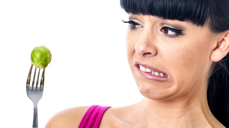 A woman looks at a Brussels sprout on a fork with disgust
