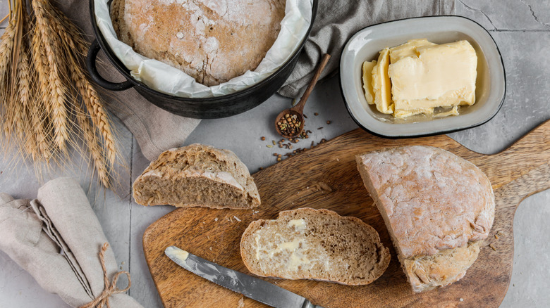 A cut sourdough loaf on a rustic chopping board with a knife, butter, and a second loaf still in its pan arranged around it