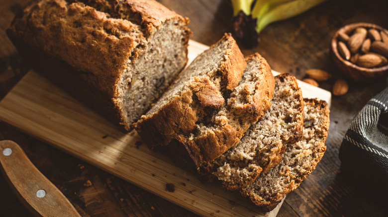 Sliced banana bread on a cutting board with nuts and fresh bananas