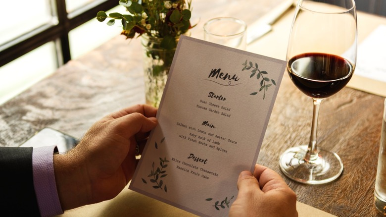 A close-up of a man holding a restaurant menu with a glass of wine nearby.