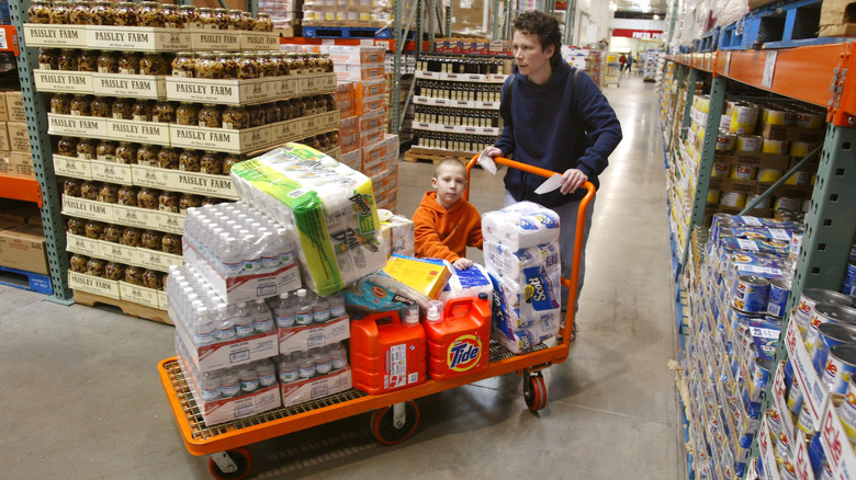 A man in Costco pushing a pallet with water, paper towels, laundry detergent, and a child riding on it