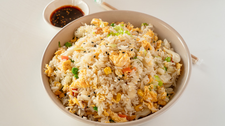 A white bowl of egg fried rice with vegetables and a side of soy sauce