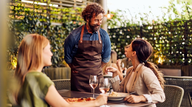 Two women out to eat together speaking to a smiling waiter wearing a brown apron.