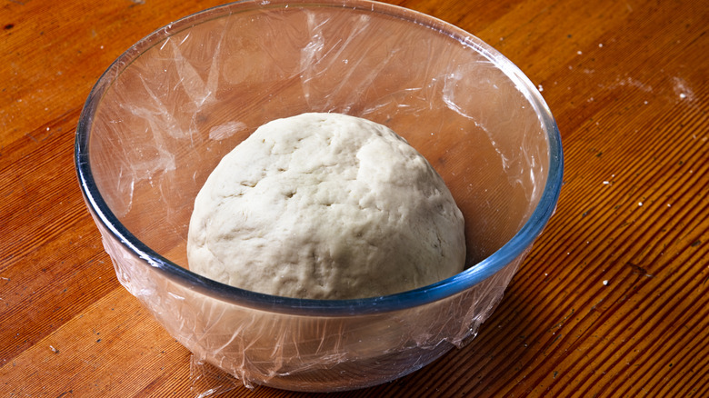 A ball of pizza dough resting in a glass bowl on a wooden surface.
