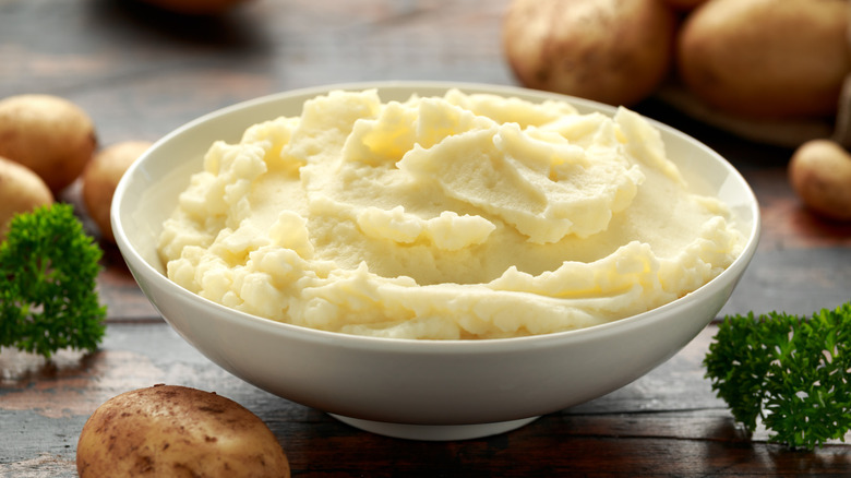 A bowl of mashed potato on a wooden table surrounded by potatoes and parsley